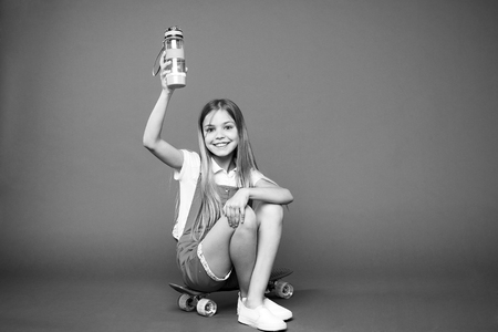 Staying hydrated. Girl happy face holds with water bottle while sit penny board violet background. Kid girl care body hydration. Active leisure and water balance. Active and healthy kid drink water.の写真素材