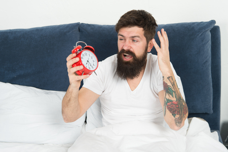 Get up with alarm clock. Overslept again. Tips for waking up early. Tips for becoming an early riser. Man bearded hipster sleepy face in bed with alarm clock. Problem with early morning awakening.の写真素材