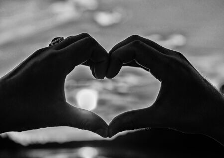 Sunset sunlight romantic atmosphere. Male hands in heart shape gesture symbol of love and romance. Heart gesture in front of sunset above river water surface, defocused. Top places for romantic dateの写真素材