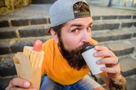 Drink something different. Caucasian hipster enjoy drinking takeaway drink with hot dog. Bearded man taking a sip of fresh coffee drink in disposable cup. Nutritional food and drinkの写真素材