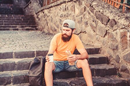 Urban lifestyle and unhealthy nutrition. Carefree hipster eat junk food while sit on stairs. Hungry man snack. Junk food. Guy eating hot dog. Man bearded enjoy quick snack. Street food so goodの写真素材