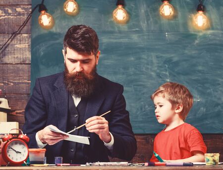 Arts lesson concept. Teacher with beard, father drawing while little son hang out near, in classroom, chalkboard on background. Child and teacher on busy face painting, drawingの写真素材