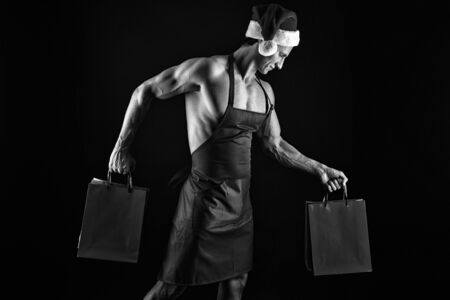 Sexy muscular man in santa hat. Christmas shopping. Present for Xmas. New year party. Santa claus man. Happy winter holidays. Red. Housewife guy with shopping bags, copy space. Shopping Bagsの写真素材