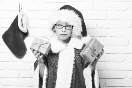 young cute santa claus boy with glasses in red sweater and new year hat with decorative christmas or xmas stocking or boot holding gifts wrapped in craft paper on white brick wall backgroundの写真素材