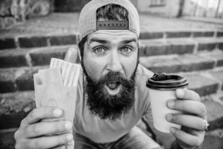 Urban lifestyle nutrition. Junk food. Carefree hipster eat junk food while sit stairs. Snack for good mood. Guy eating hot dog. Street food concept. Man bearded eat tasty sausage and drink paper cupの写真素材