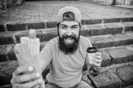 Snack for good mood. Guy eating hot dog. Man bearded eat tasty sausage and drink paper cup. Street food concept. Urban lifestyle nutrition. Junk food. Carefree hipster eat junk food while sit stairsの写真素材
