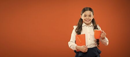 A healthy taste. Cute girl with tea cup and note book on orange background. Little schoolchild drinking tea for breakfast. Small kid enjoying morning tea. Child having tea break in school, copy spaceの写真素材
