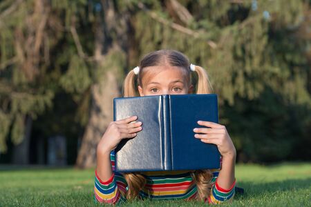 Dont judge book by its cover. Adorable small child cover face with book. Cute little girl read book on green grass. Great eye for good book.の写真素材