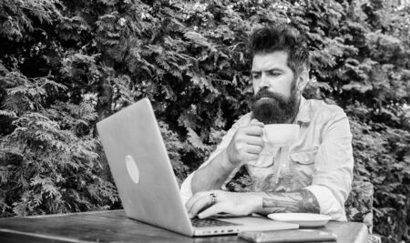 Work and play give him rest and energy. Bearded man doing his work online. Hipster drinking tea and using computer work station outdoor. Working globally through distance workの写真素材