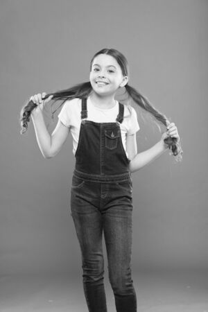 Feeling good fabulous hair. Adorable girl holding ponytails hair on orange background. Small cute girl with long brunette hair wearing casual style. Little hair model with fashion lookの写真素材