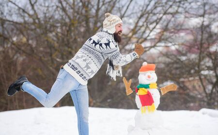 Man cheerful bearded hipster knitted hat and warm gloves play with snowman snow outdoors. Have fun winter day. Let it snow. Christmas holidays. Active lifestyle. Snow games. Leisure on fresh airの写真素材