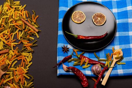 delicious colorful Italian pasta ceramic plate with wooden spoon , chili pepper, orange, garlic, star anise and cinnamon white and blue plaid fabric or checkered cloth on dark background, side viewの写真素材