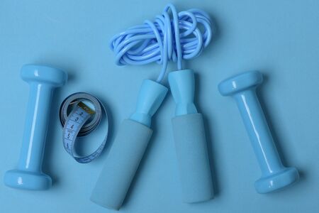 Health regime and fitness symbols. Dumbbells and skipping rope in cyan color on blue background, top view. Healthy lifestyle and sports concept. Jump rope and barbells next to rolled measure tapeの写真素材