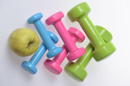 Health regime and fitness symbols. Dumbbells near green apple on white background. Healthy lifestyle and sports concept. Apple fruit and pink, green and blue barbells making pattern, top viewの写真素材