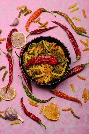 delicious colorful Italian pasta in ceramic plate with spoon , chili pepper, orange, garlic on pink textured background, top viewの写真素材