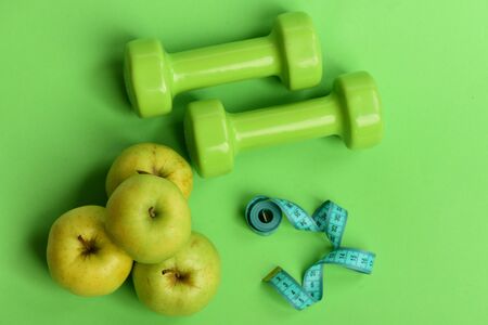 Tape measure in cyan color by barbells and juicy apples, topview. Healthy lifestyle symbols. Dumbbells in bright color, rolled measure tape and fruit on green background. Diet and sport regime conceptの写真素材