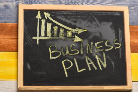 Chalkboard with inscription written in yellow. Simple business decrease graph. Business plan concept. Blackboard with business chart on colorful wooden background.の写真素材