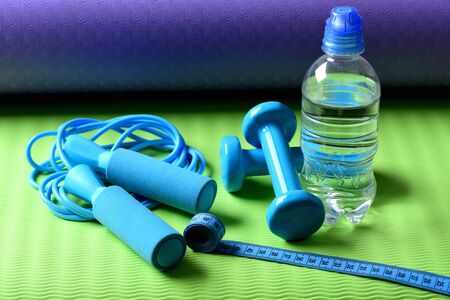 Workout and refreshment concept. Skipping rope, cyan dumbbells and tape near water bottle on green and purple background. Bottle or water near jump rope and barbells on yoga mat. Sports and healthの写真素材