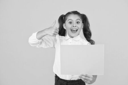 This is genius. Happy girl genius holding blank paper sheet on yellow background. Genius child giving thumbs up gesture for empty poster. Liking her genius idea, copy spaceの写真素材