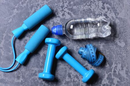 Sports and health idea. Dumbbells, cyan skipping rope, water bottle and measuring tape on grey texture background. Workout and measurement concept. Water and tape near jump rope and plastic barbellsの写真素材