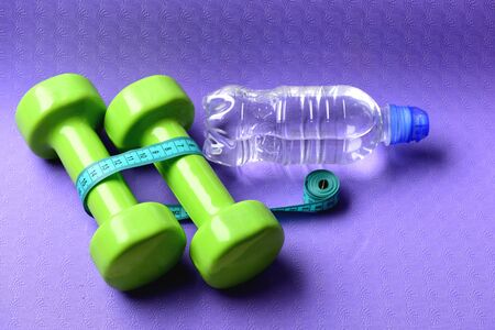 Healthy shape and sport concept. Barbells next to roll of tape measure and water bottle. Shaping and fitness equipment. Dumbbells and measure tape in cyan blue color on purple backgroundの写真素材