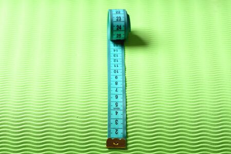 Measuring tape roll in cyan color on green texture background. Sports and healthy lifestyle concept. Shaping and fitness equipment, close up. Flexible ruler in blue lying on yoga mat, selective focusの写真素材