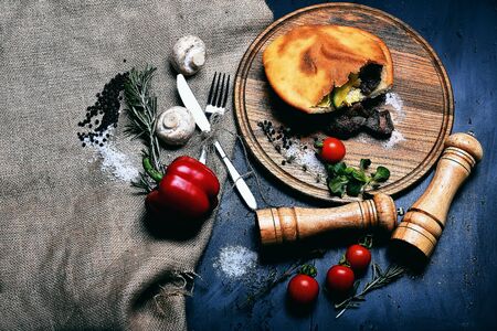 calzone or pie with meat, fresh red cherry tomato, sweet pepper, mushroom, rosemary, thyme, corn salad, salt on wooden tray, pepperbox, saltcellar, silver fork, knife on grey background with burlapの写真素材