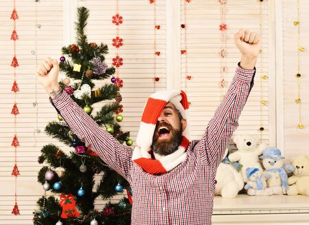 Man with beard puts hands up. Celebration and partying concept. Guy in red and white scarf wrapped around his head standing by Christmas tree. Santa Claus with excited face on wooden wall backgroundの写真素材