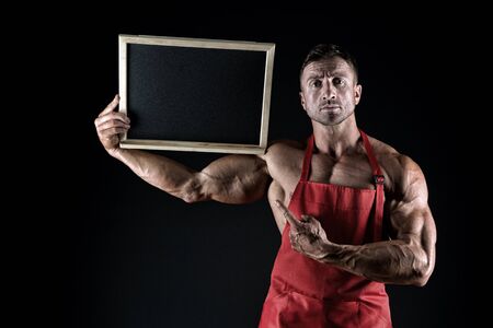 sexy and muscular chef show blackboard. copy space. athletic bodybuilder in cook apron. housekeeper pointing finger on board. place for your advertisement. healthy menu here. beat food and coffeeの写真素材