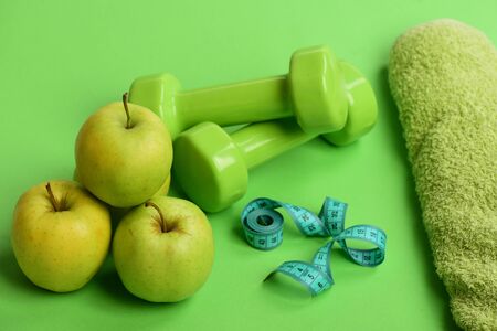 Barbells by green apple. Healthy lifestyle and low calorie food concept. Sports and healthy regime equipment. Dumbbells in bright green color, twisted measure tape, towel and fruit on green backgroundの写真素材