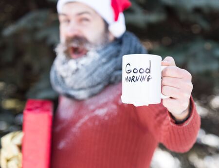 White cup with good morning text in hand of handsome happy man in santa claus hat, bearded hipster...
