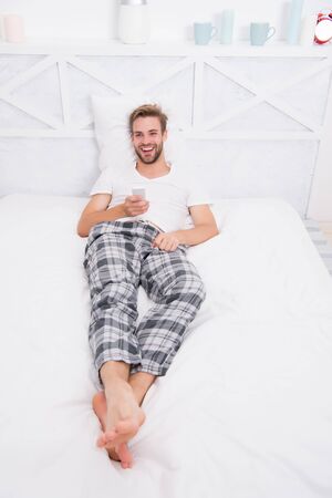 being in good mood. lazy sunday weekend at home. watch morning show. man watch tv. change channel by remote control in bed. happy man in pajama with remote control. guy watching television in bedroomの写真素材