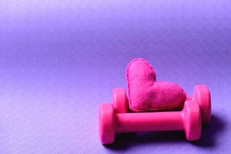 Barbells and pink love symbol, close up. Dumbbells made of pink plastic near soft toy heart on purple texture background, copy space. Healthy shape concept. Love of sports and fitness ideaの写真素材