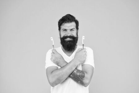 Guy hold two toothbrushes. Hipster happy smiling face cares hygiene. Brush teeth. Teeth whitening procedure. Ultrasonic teeth cleaning technology. Bearded man holds toothbrushes yellow backgroundの写真素材