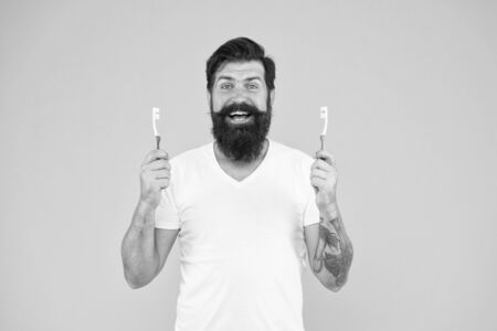 Bearded man holds toothbrushes yellow background. Guy holds two toothbrushes. Hipster happy smiling face cares hygiene. Brush teeth concept. Teeth hygiene. Ultrasonic teeth cleaning technologyの写真素材