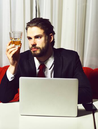 bearded man, businessman holding glass of whiskey works at laptop