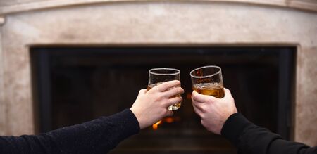 Glasses with whisky in male hands. Hands clinking whisky glasses on fireplace background. Male hands with drinks in glasses at home by fireplace. Men cheers with whisky in glasses near fireplaceの写真素材