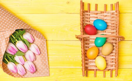 Colorful bright eggs and bouquet fresh tulip flowers on yellow background top view. Spring holiday....