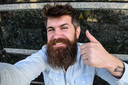 Hipster, tourist with tousled hair and long beard looking at camera, taking selfie photo. Selfie photo concept. Man, with beard on cheerful face shows thumb up gesture, black marble background.の写真素材