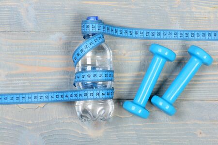 Water, measuring tape dumbbells, top view. Workout and losing weight concept. Tools for healthy and active lifestyle. Sports equipment in cyan blue and bottle lay on wooden vintage background.の写真素材