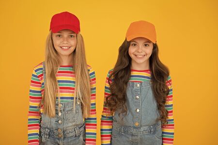 Hip hop girls. Little girls smiling on yellow background. Happy small girls wearing baseball caps in casual style. Adorable caucasian girls with long loose hairの写真素材