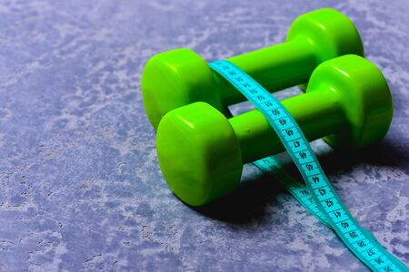 Dumbbells in green color and cyan measuring tape around them on grey texture background. Workout and measurement concept. Measuring tape around plastic barbells. Sports and healthy training ideaの写真素材
