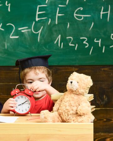 Cute boy in graduation cap playing with clock. Little kid learning how to tell the time. Blond child sitting at the desk in the kindergarten.の写真素材