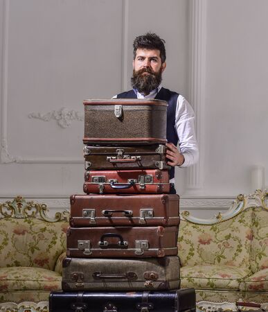 Macho elegant on strict face standing near pile of vintage suitcase. Luggage and travelling concept. Man, traveller with beard and mustache packing luggage before trip, luxury interior background.の写真素材
