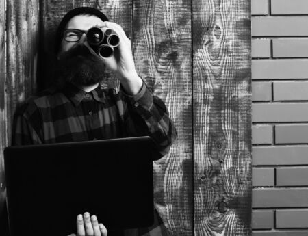 Bearded man, long beard. Brutal caucasian serious unshaven hipster holding laptop and craft paper rolls in red black checkered shirt with hat and glasses on brown vintage wooden studio backgroundの写真素材