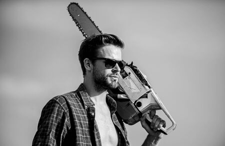 Get stylish haircut. Barbershop concept. Masculinity concept. Handsome man with chainsaw blue sky background. Dangerous job. Powerful chainsaw. Lumberjack hold chainsaw. Gardener lumberjack equipmentの写真素材