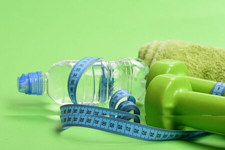 Healthy regime symbols. Tape measure in cyan color near barbells and water bottle. Athletics and weight loss concept. Dumbbells in bright green color, towel and tape around bottle on green backgroundの写真素材