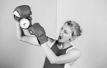 Establishing the clock time. Cute boxer with angry look fighting with alarm clock. Emotional woman holding clock in boxing gloves. Sportswoman punching a clock. Its time for boxing trainingの写真素材