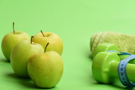 Barbells by juicy green apples. Sports regime equipment, copy space. Dumbbells in bright green color, measure tape, towel and fruit on green background. Athletics and weight loss conceptの写真素材