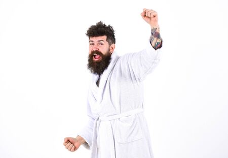 Perfect morning concept. Happy guy saluting with his raised arm. Cheerful man in white bathrobe standing in white roomの写真素材